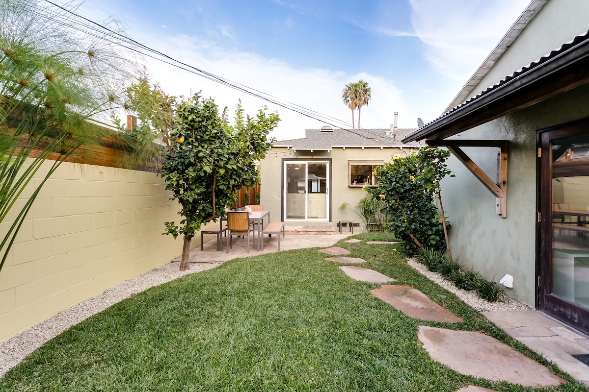 A spacious backyard features a neatly manicured lawn, bordered by plants and citrus trees. A stone pathway leads to a dining table set on a patio, with a light-colored building in the background and clear skies above.