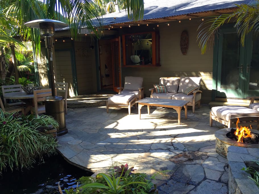 Back yard stone patio offers tranquil setting in lush tropical landscape with koi pond and water falls, cushioned teak lounge furniture, dining, fire pit, grill, and bamboo pavilion.
