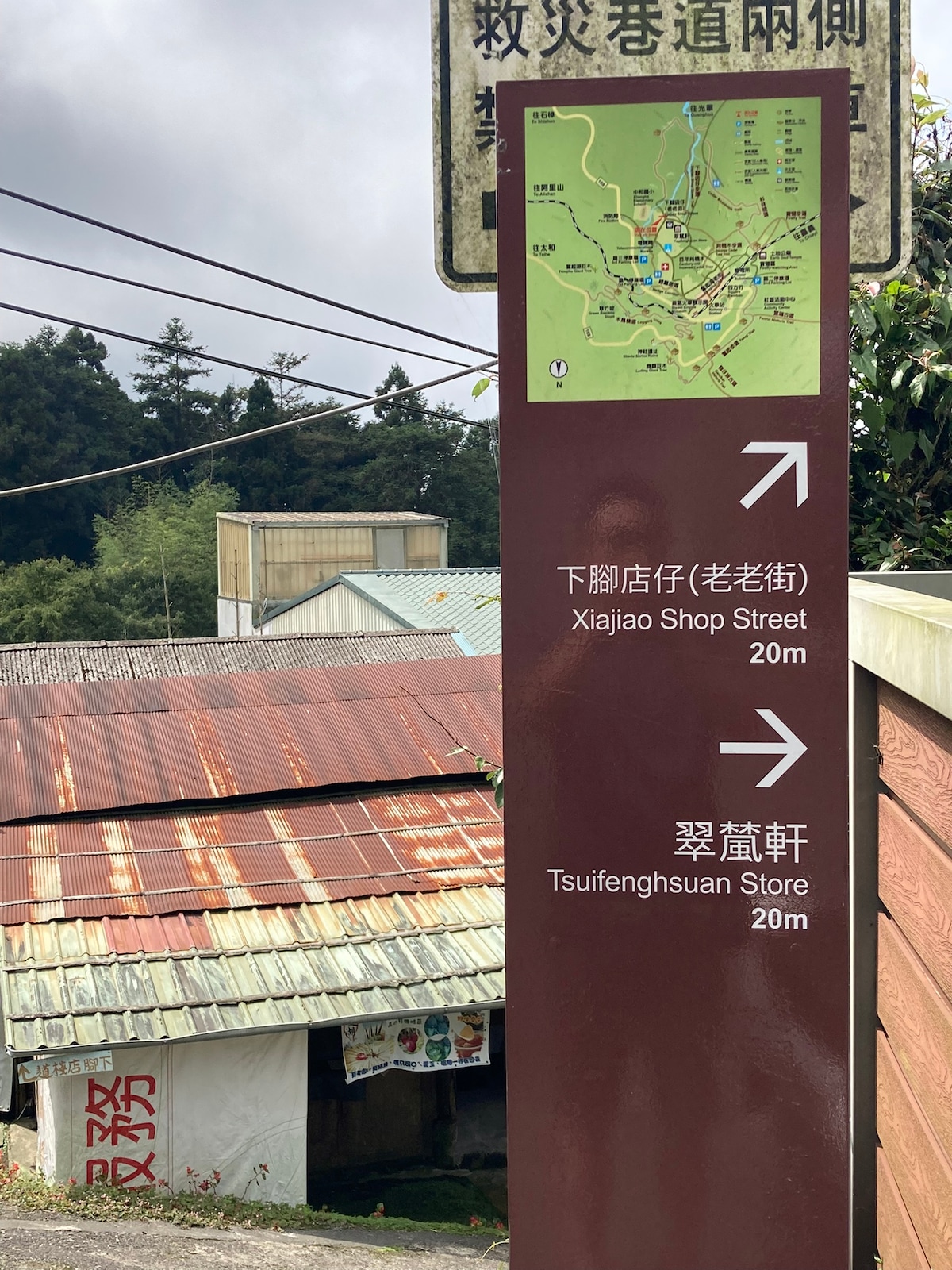 A directional sign displays maps and distances to nearby locations, including Xiajiao Shop Street and Tsufenghsuan Store, both 20 meters away. The background features a collection of buildings with varied roofs, including a sloped metal structure and a partially visible wall with text.