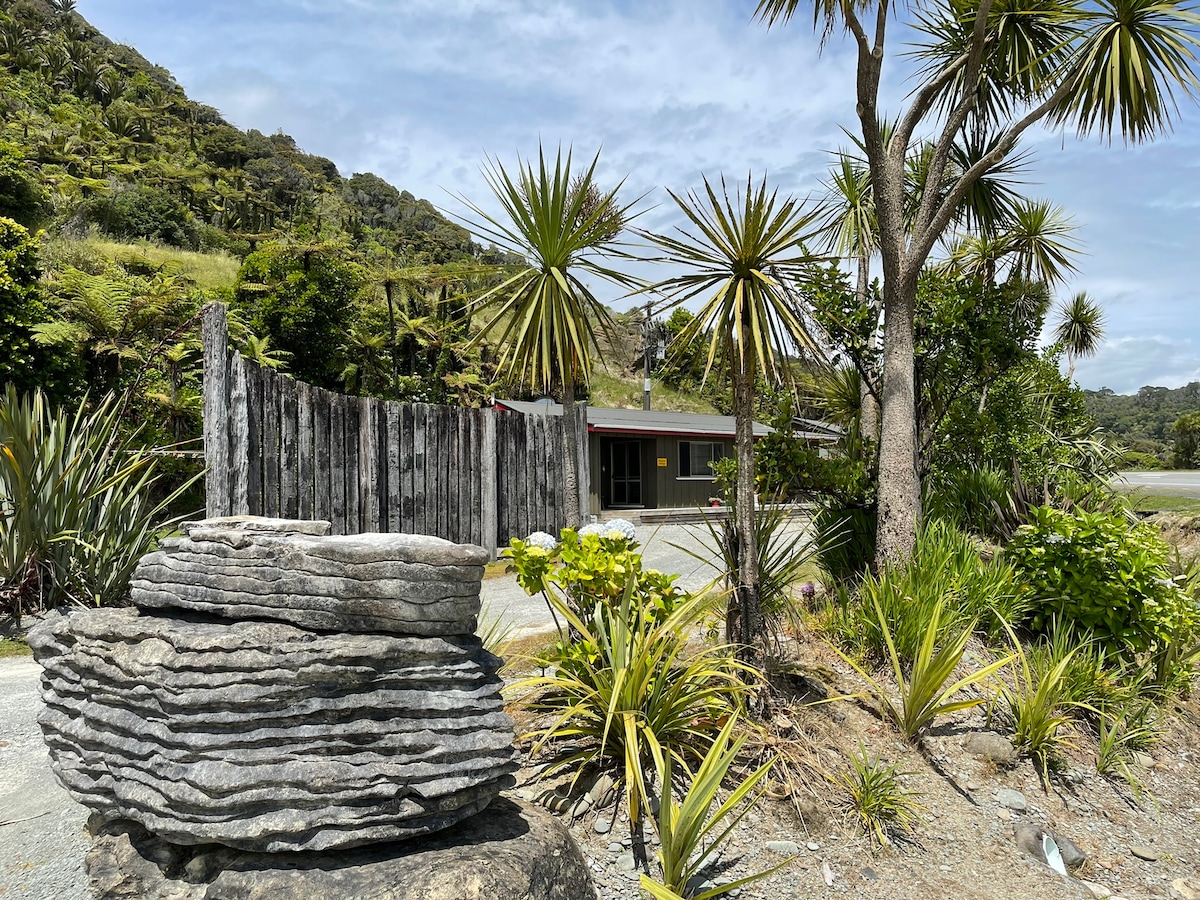 A natural stone feature is framed by lush tropical plants and palm trees, with the cottage entrance visible in the background. The wooden fence adds texture to the scene, and the landscape features rolling greenery, creating a sense of privacy and tranquility.