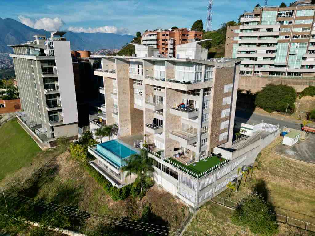 The modern apartment building features a distinctive architectural design, surrounded by greenery. A rooftop terrace is visible, highlighting outdoor space with a pool below. Scenic views of the mountains and urban landscape can be observed in the background.