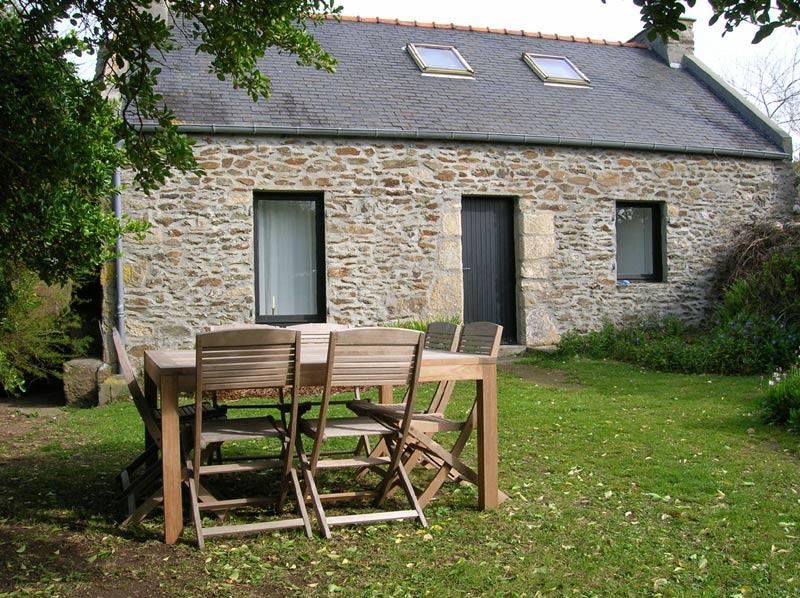A traditional stone house is showcased, featuring two skylights and a black door framed by two windows. A wooden dining table with six chairs is positioned on a grassy area, surrounded by greenery. The setting reflects a peaceful outdoor environment.