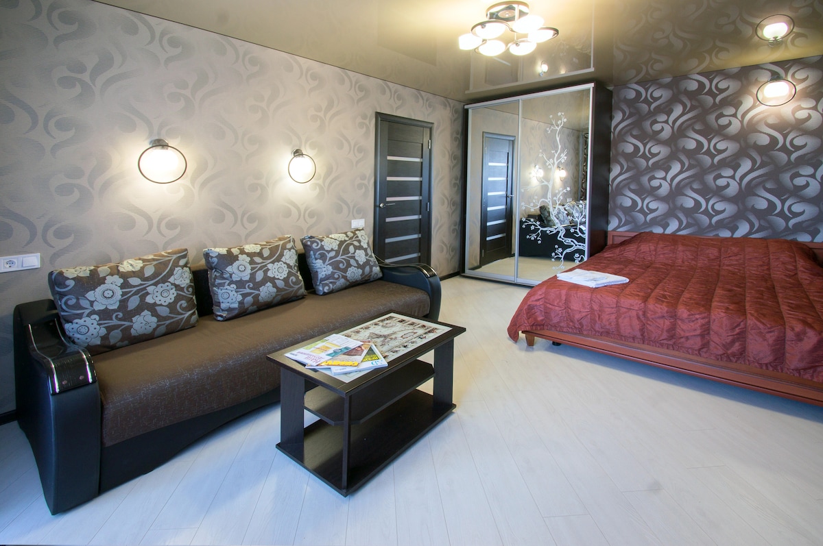The living area features a dark sofa adorned with floral patterns, paired with a sleek coffee table. A spacious bed with a deep red cover is positioned against a patterned accent wall. A sliding mirrored wardrobe reflects the soft lighting from the decorative fixtures.