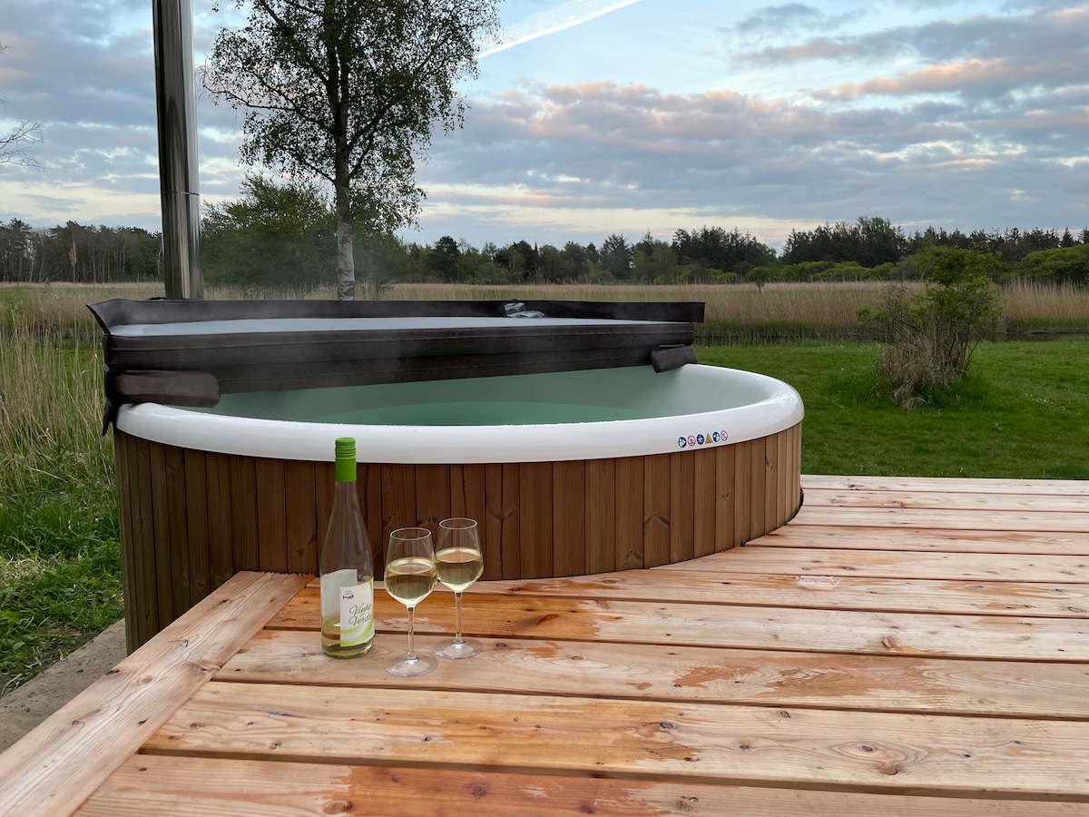 A wooden deck features a hot tub, steam rising gently from the surface. Glasses of white wine sit on the table, complemented by a bottle nearby. The surrounding landscape of tall grass and trees creates a peaceful setting.