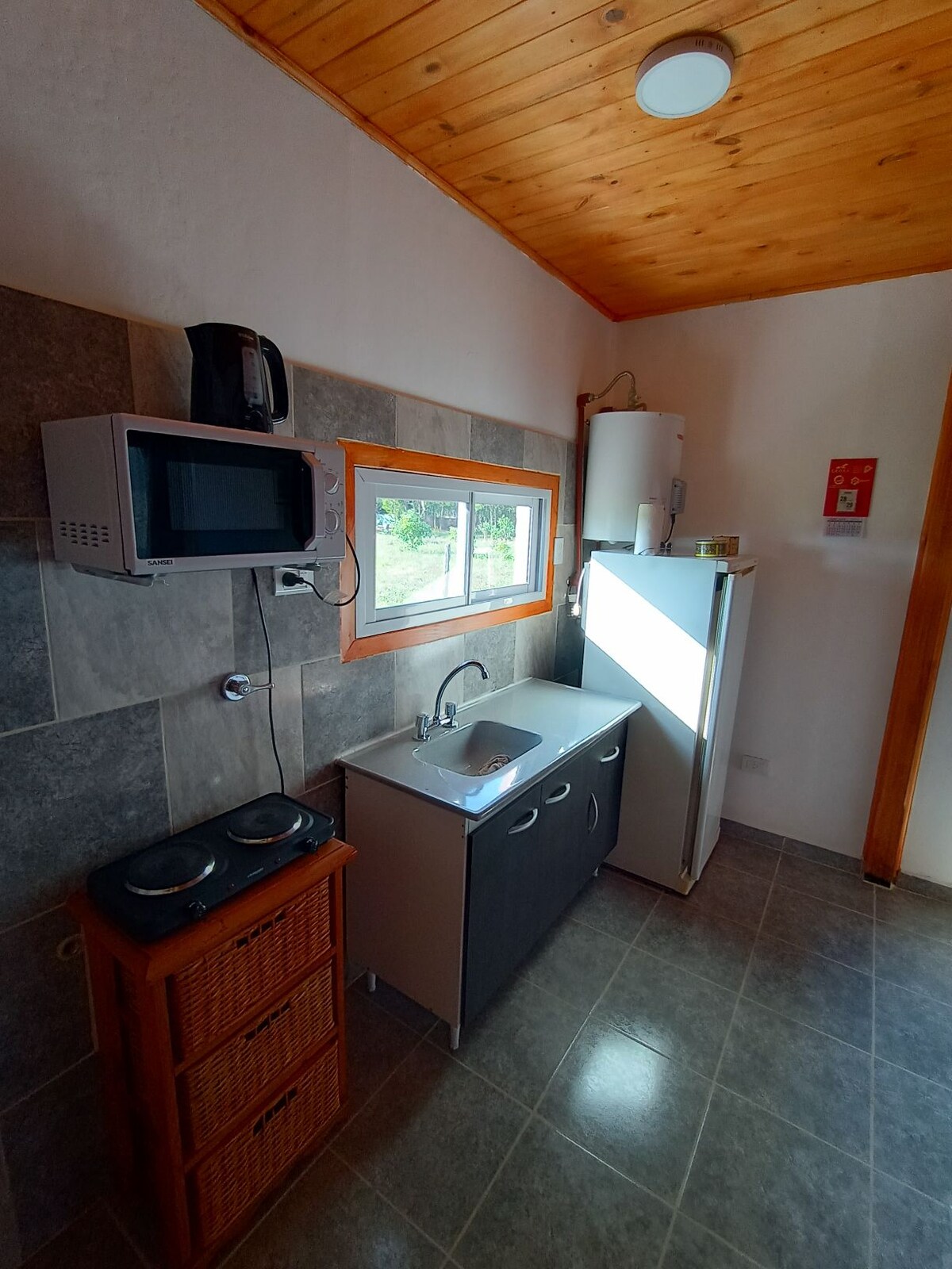 A functional kitchen area is presented with modern appliances, including a microwave and refrigerator. A compact sink is positioned under a bright window, allowing natural light to enhance the space. Cupboard storage is complemented by a woven basket unit, providing an organized feel.