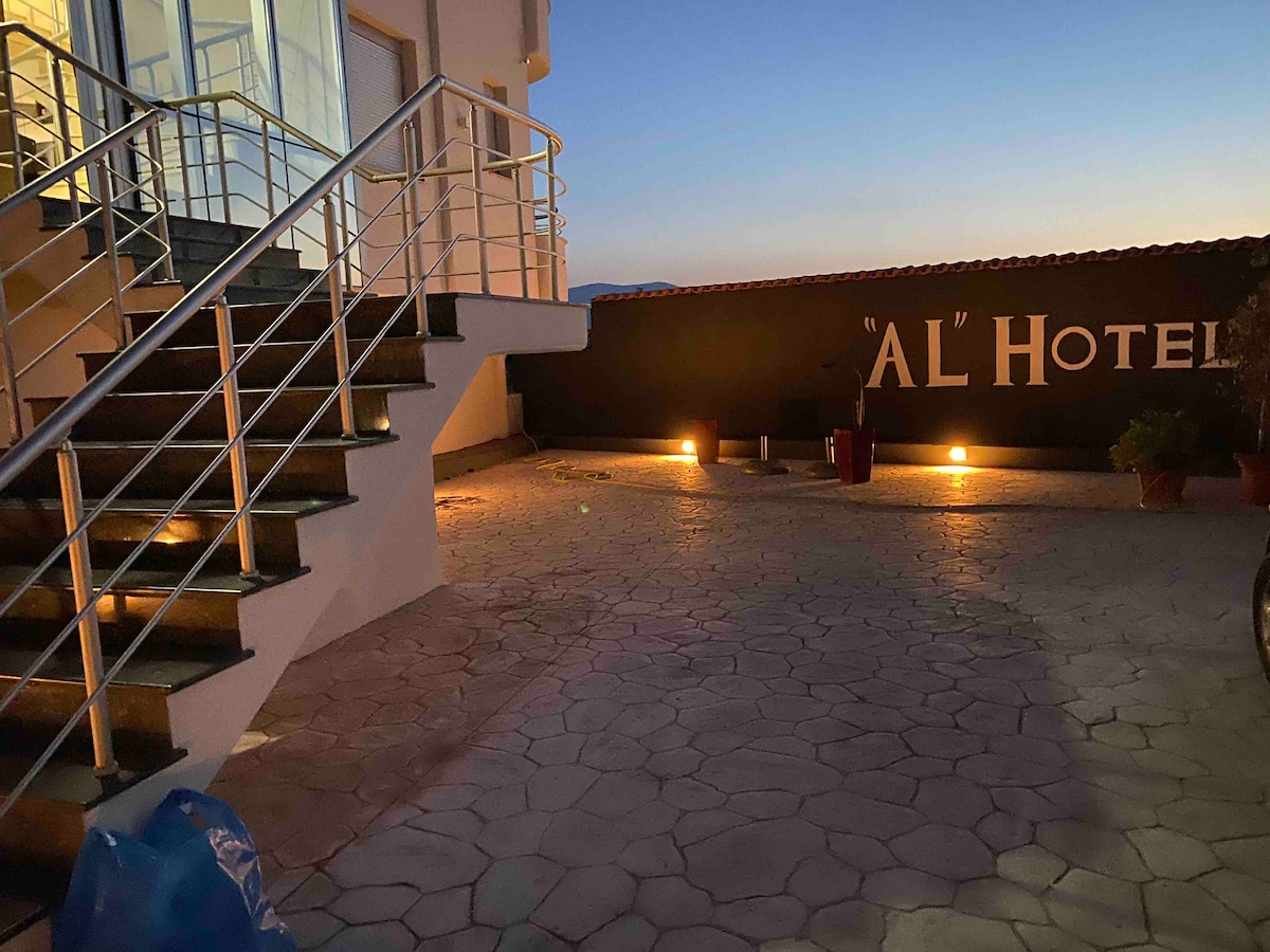 An exterior view of the hotel entrance is shown, featuring a staircase with a stainless steel railing. The area is illuminated by soft lighting, accentuating the textured stone pavement. The hotel name, 'AL,' is displayed prominently on a wall, creating a welcoming atmosphere.