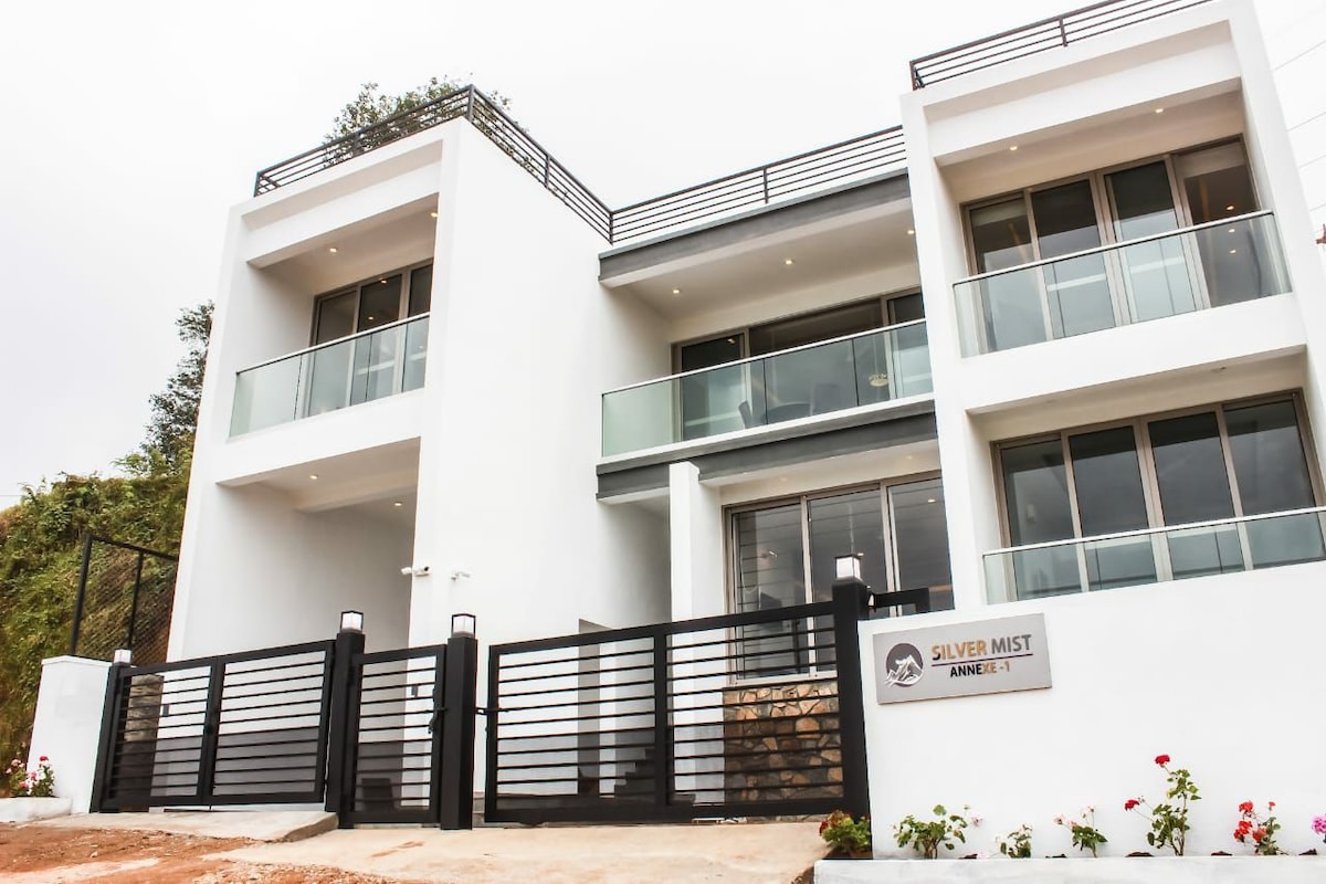 The exterior of Silver Mist Annexe I features a contemporary design with clean lines and a color palette of white and gray. Glass balconies extend from the upper floors, enhancing natural light. A modern gate provides access to the property, framed by well-kept landscaping.