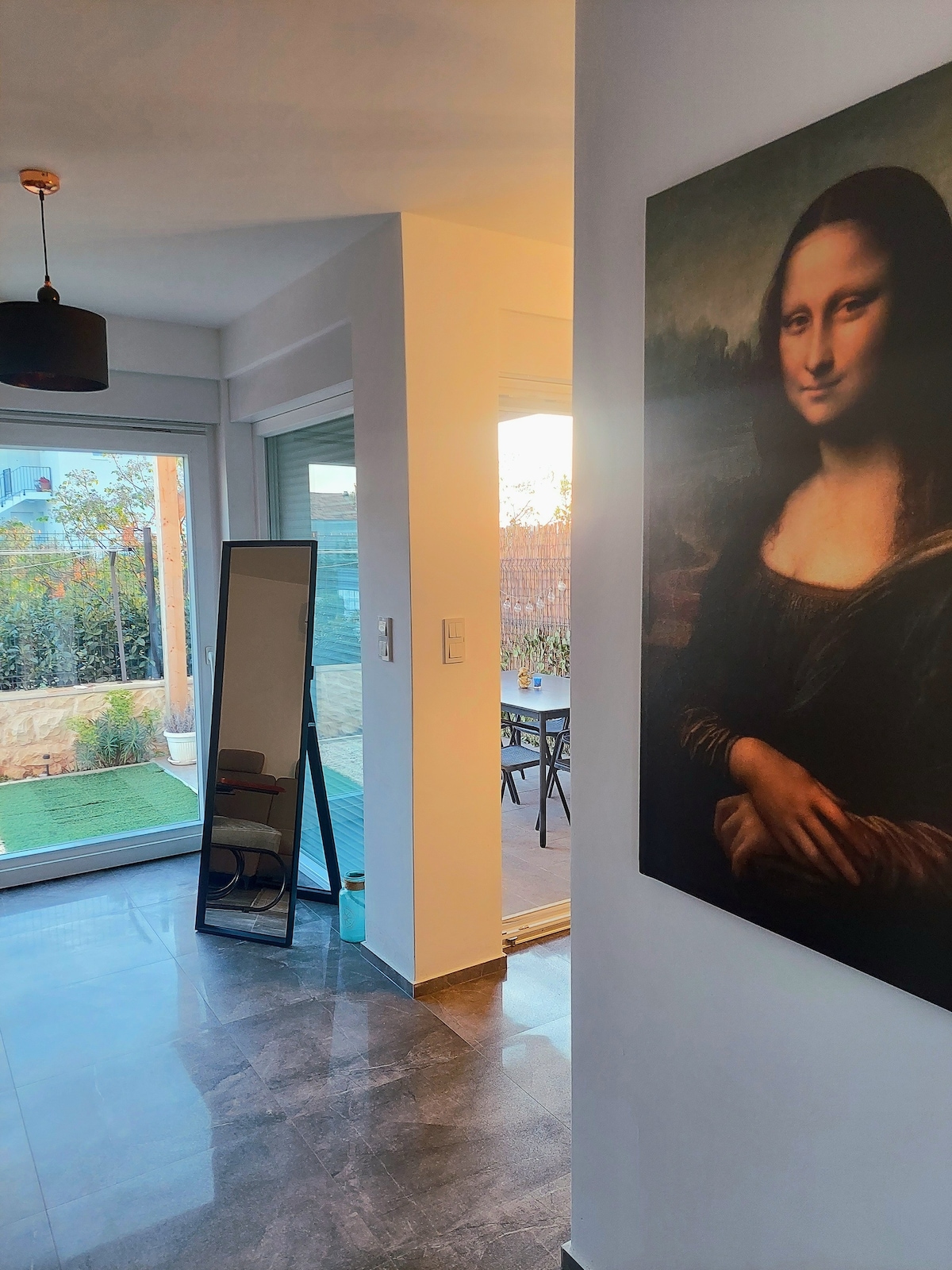 A hallway features a full-length mirror and a large artwork of a woman. Light filters in through sliding glass doors, leading to an outdoor area with a small table and chairs visible beyond. The floor is covered in sleek, dark tiles, complementing the bright walls.