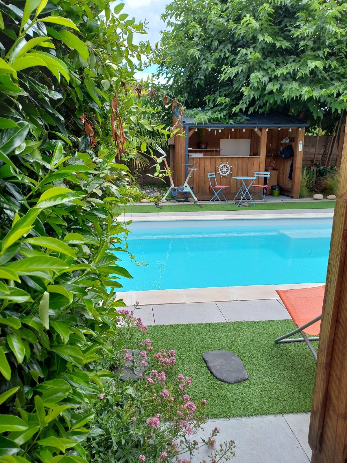 A serene view of the outdoor swimming pool is showcased, framed by lush greenery. A wooden gazebo with seating is visible in the background, along with two blue chairs set near the pool. Bright orange lounge chairs rest on the patio area, enhancing the tranquil setting.