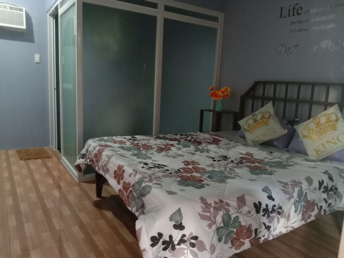 A spacious sleeping area is complemented by floral bedding spread across a large bed. Light-colored walls and a glass-enclosed bathroom contribute to a fresh ambiance. A small decorative rug is positioned near the bed, and an air conditioning unit is installed on the wall.