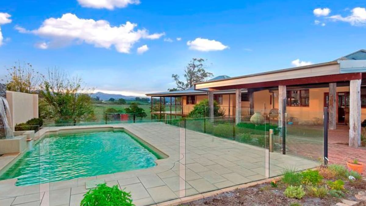 A large outdoor area features a rectangular swimming pool with a green-tinted surface. Surrounding the pool are paved patios and landscaped gardens with low shrubs, providing a clear view of the rolling hills and distant mountains.
