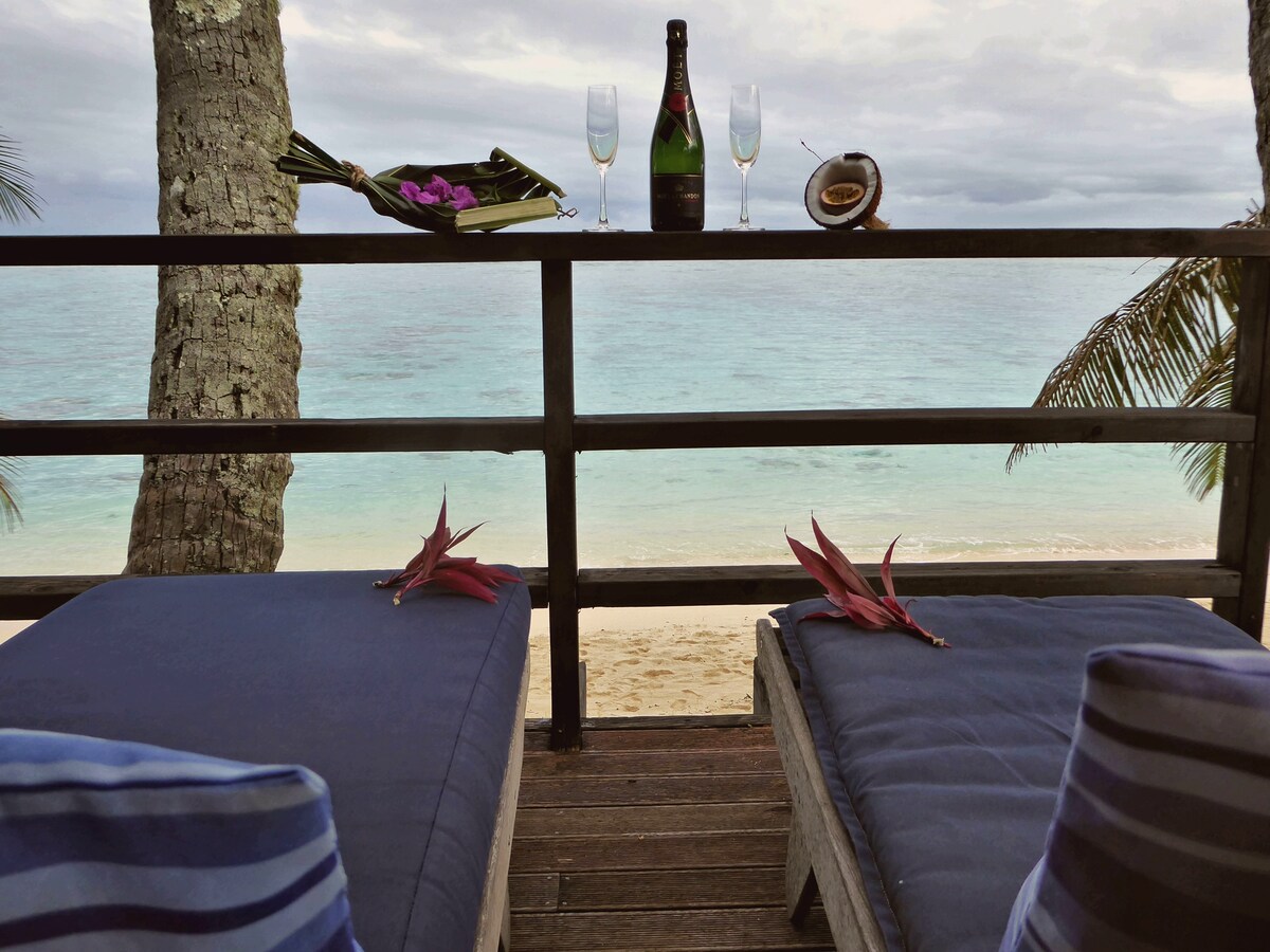 A wooden deck offers two loungers adorned with decorative cushions, overlooking a sandy beach and calm turquoise waters. A table displays two champagne flutes, a bottle of champagne, and a tasteful arrangement of tropical foliage, creating a serene and inviting atmosphere.