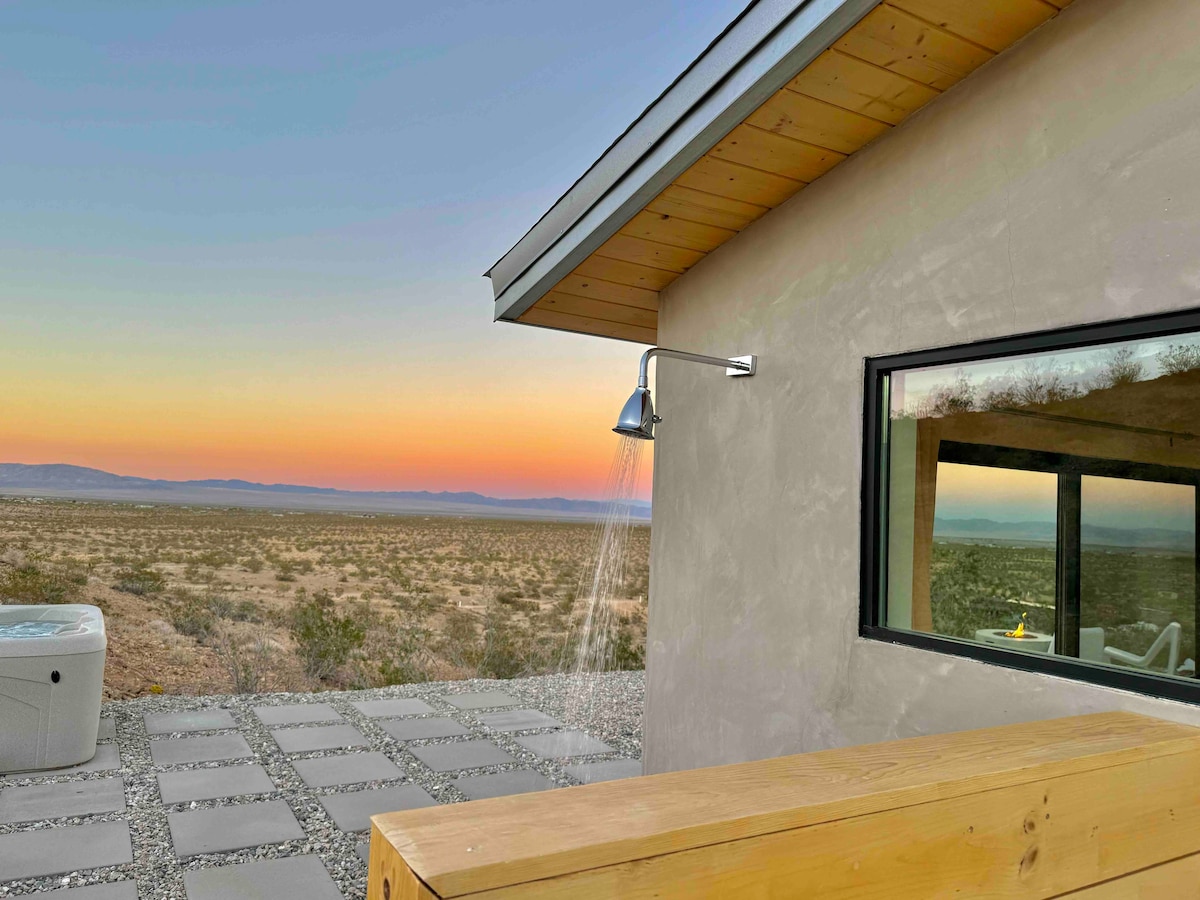 An outdoor shower is positioned alongside a modern structure, featuring sleek lines and a minimalist design. A soft sunset glow casts warm colors across the expansive desert landscape, visible through large glass windows. A hot tub sits nearby, enhancing the serene atmosphere.
