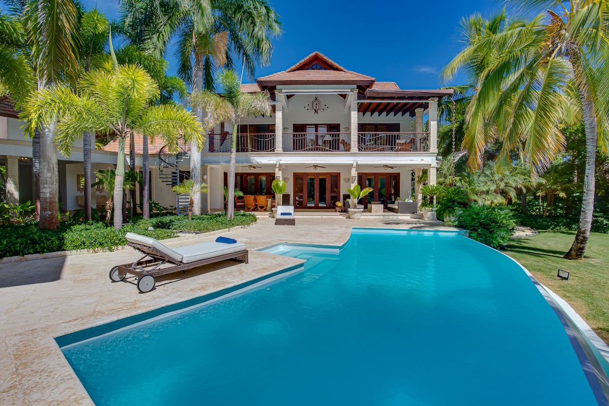 A spacious pool terrace is framed by lush palm trees, with a semi-circular pool inviting relaxation. The two-story villa stands in the background, featuring expansive balconies and large glass doors that connect indoor and outdoor living spaces. Sun loungers are arranged beside the pool.