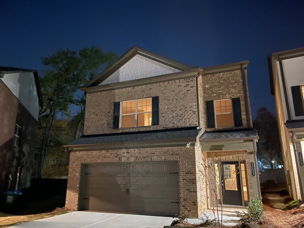 The exterior of a newly developed two-story home is visible at night, showcasing brick walls and dark shutters. Illuminated windows create a welcoming glow, while a two-car garage is situated at the front. Nearby landscaping includes small plants and a clear driveway.