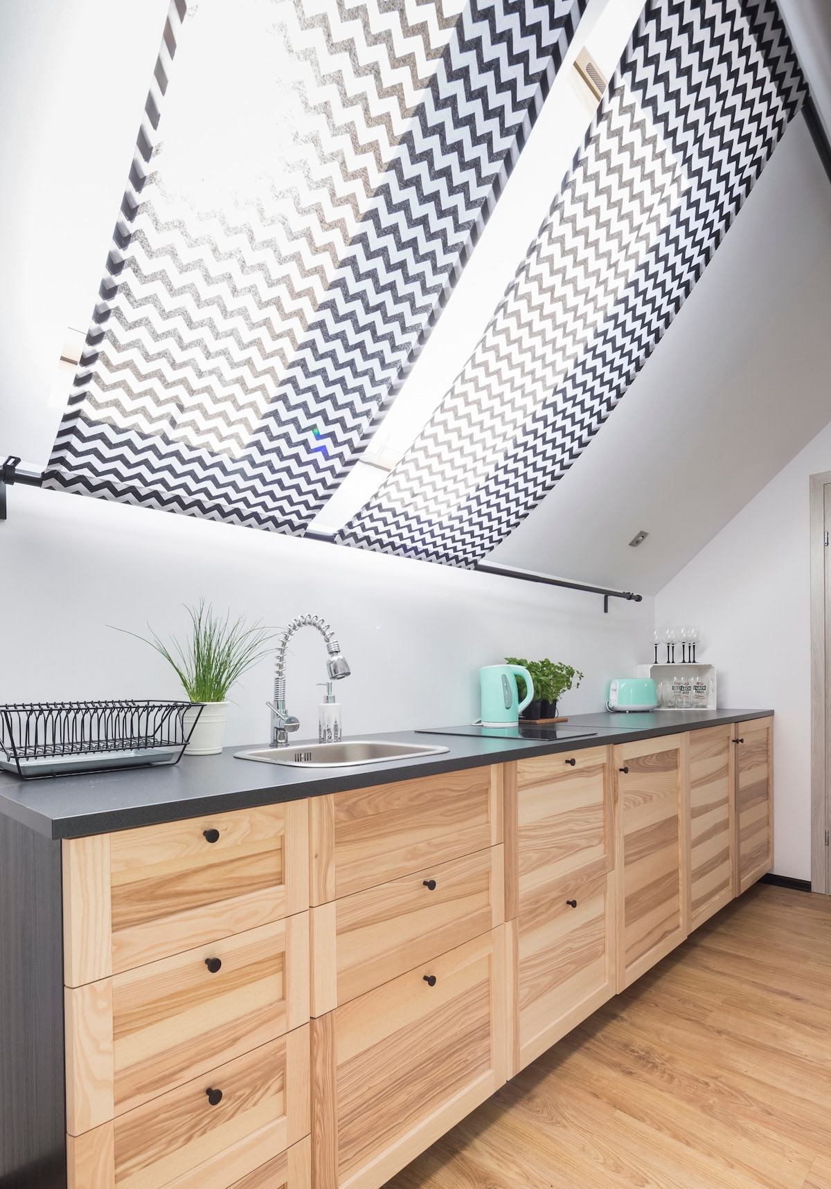 A functional kitchen area is highlighted by modern cabinetry with wood finishes, complemented by a dark countertop. A sink is positioned under a large, patterned window covering that allows natural light to fill the space. A small plant and a kitchen appliance set add practicality.