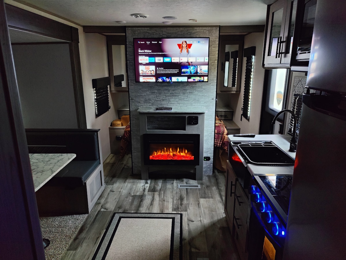 A modern camper interior is displayed, featuring a cozy seating area with a table and comfortable seating adjacent to a stylish kitchen. An electric fireplace adds warmth, while a large television provides entertainment. Ample natural light filters through the windows with sleek blinds.