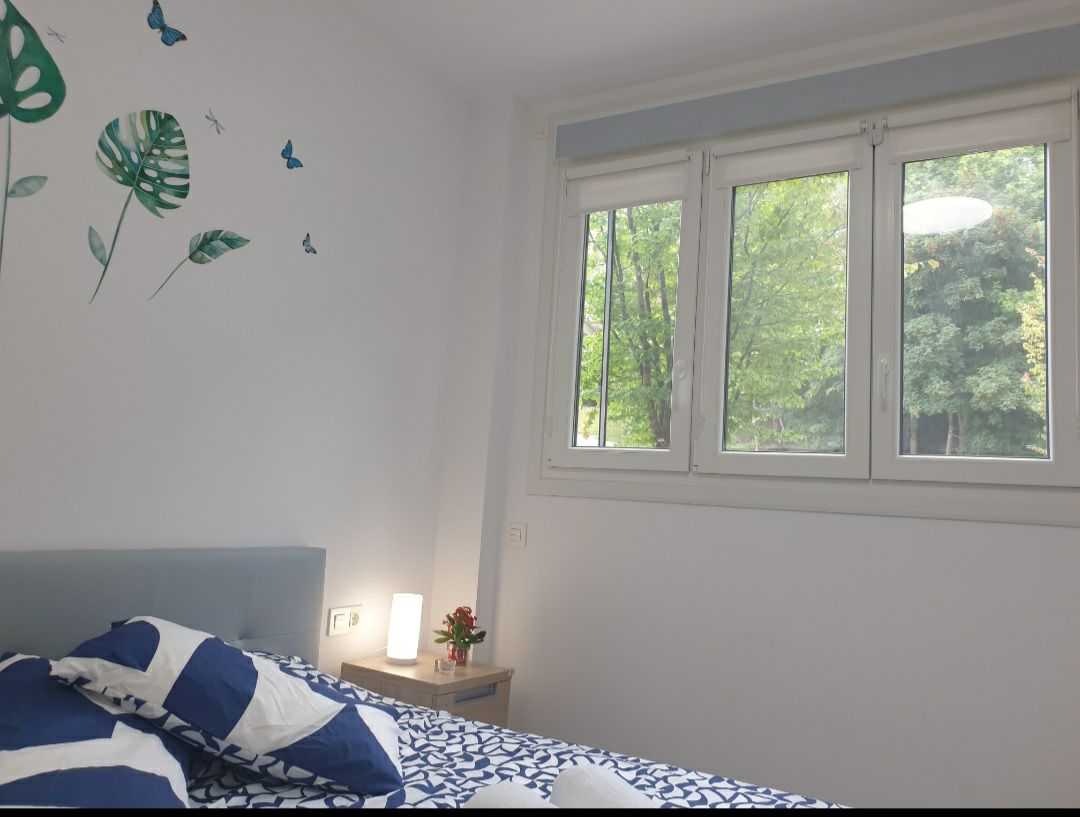A bright bedroom features a spacious bed dressed in a blue and white patterned coverlet. Windows with multiple panes offer views of the greenery outside, allowing natural light to fill the space. A small lamp and a decorative plant are positioned on a bedside table.