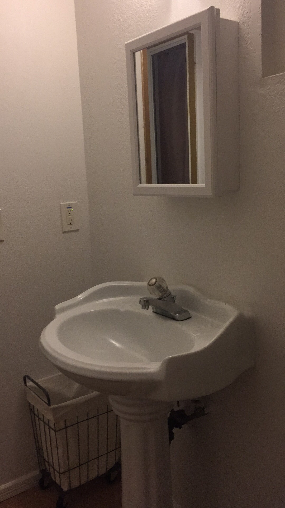 A small, well-maintained bathroom features a pedestal sink with a single faucet. Above the sink, a rectangular mirror is mounted on the wall. A wire basket is positioned nearby, providing a space for additional toiletries or towels. Soft lighting enhances the functional space.