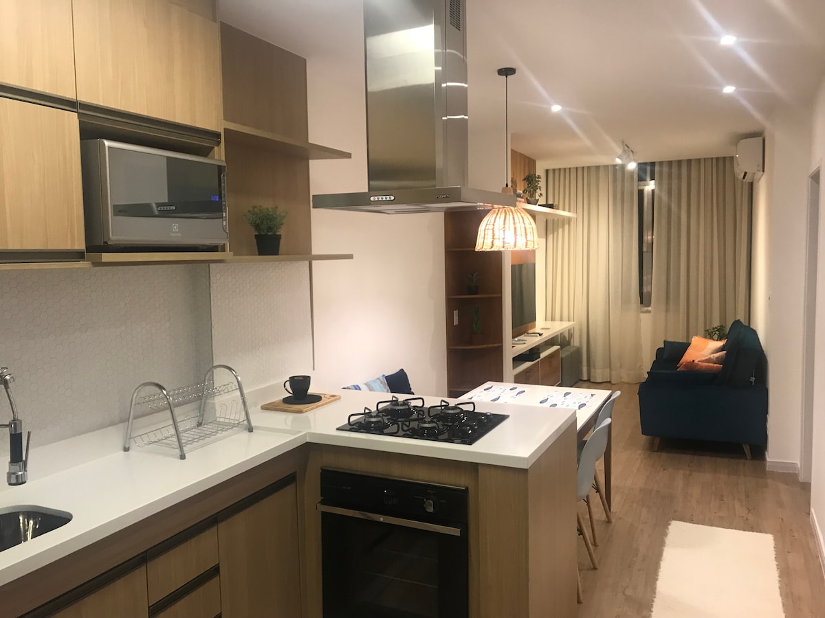 A modern kitchen features sleek wooden cabinets and a gas stove with a built-in oven. A dining area with a polished table is visible in the foreground, while a cozy living space with a blue sofa and a TV can be seen in the background, illuminated by soft lighting.