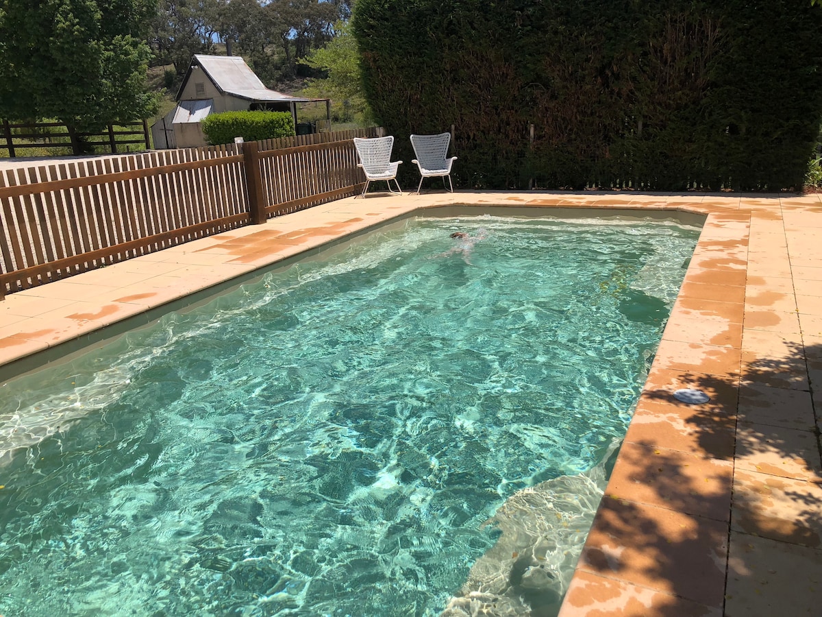 A private swimming pool is surrounded by a paved terrace, featuring clear water reflecting sunlight. Two white lounge chairs are positioned nearby, offering a space for relaxation. A wooden fence and well-maintained hedge provide a sense of seclusion.