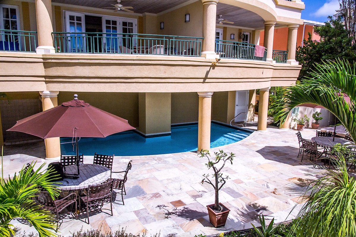 An outdoor area features a small, private pool bordered by a stone patio. Several dining chairs and tables are arranged under a shaded umbrella. Lush greenery and a potted plant add a tropical touch to the serene space.