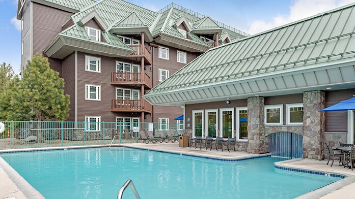 A swimming pool is situated in the foreground, featuring clear blue water. Surrounding the pool, lounge chairs are arranged neatly, alongside a building with a distinctive green roof and multiple balconies. The area is enclosed by a green fence, offering a sense of privacy.