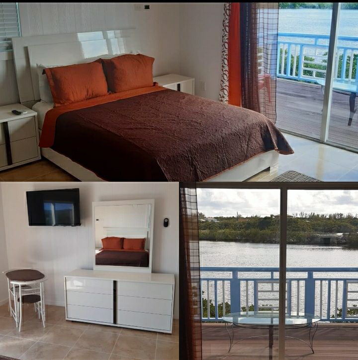 A bright sleeping area features a spacious bed with an earthy-toned coverlet and coordinating pillows. Natural light filters through large windows, revealing a scenic view of the water. A mirror and dresser complement the room, while a small table and chairs are positioned on the balcony.