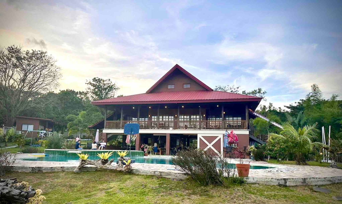 A spacious two-story house features a red roof and a large covered porch. The surrounding area includes well-maintained gardens, a sparkling pool, and a variety of tropical plants. The outdoor space is designed for relaxation and gatherings, inviting friends and families to enjoy time together.