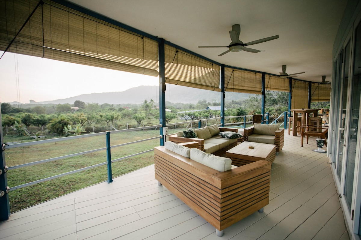 A spacious outdoor decking area features wooden furniture arranged for relaxation. Soft cushions are placed on the seating, creating a comfortable space to enjoy the views. The background includes a lush landscape with greenery and distant mountains, complemented by ceiling fans for airflow.