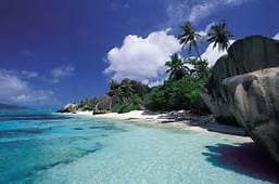 A serene beach scene features soft white sand meeting clear turquoise waters. Lush greenery is visible in the background, complemented by scattered palm trees and unique rock formations, under a bright blue sky dotted with fluffy clouds.