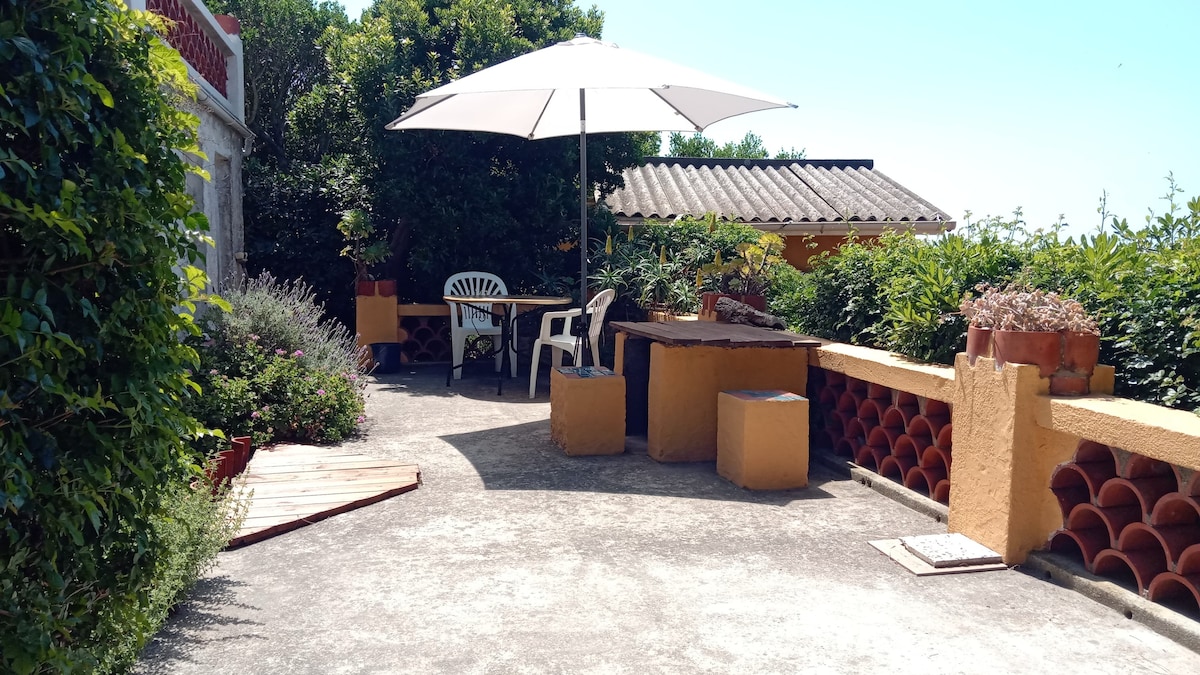 A spacious terrace is furnished with a round table and chairs, providing shaded seating under a large umbrella. Surrounding greenery and colorful planters contribute to a tranquil outdoor setting.