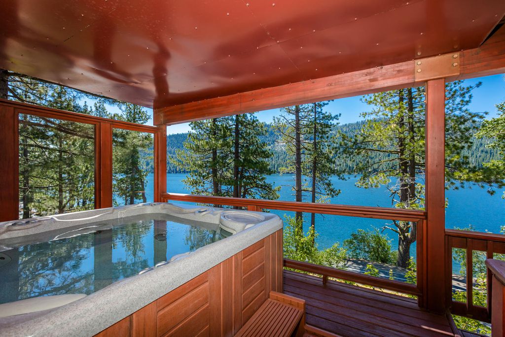 A hot tub is situated on a wooden deck, offering serene views of the blue water and surrounding pine trees. The enclosure features large windows that allow natural light and a stunning view of the landscape beyond.