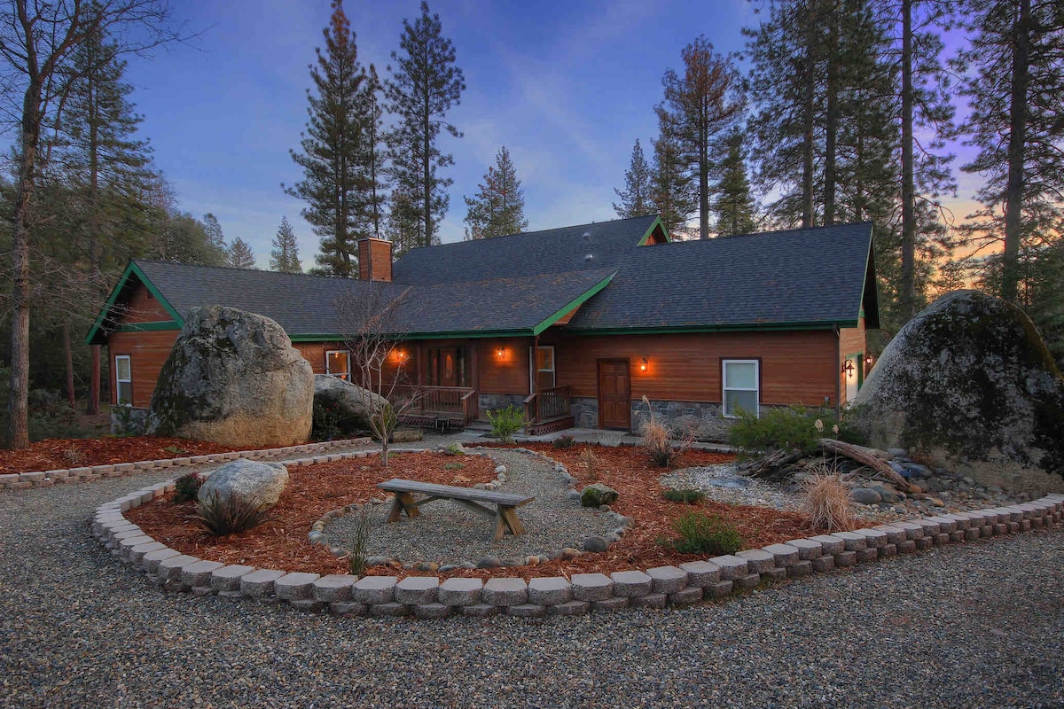 A spacious exterior view of a rustic wooden house is framed by tall pine trees and granite boulders. A stone pathway leads to a circular seating area with a picnic table, surrounded by decorative landscaping and gravel, all set against a soft twilight sky.