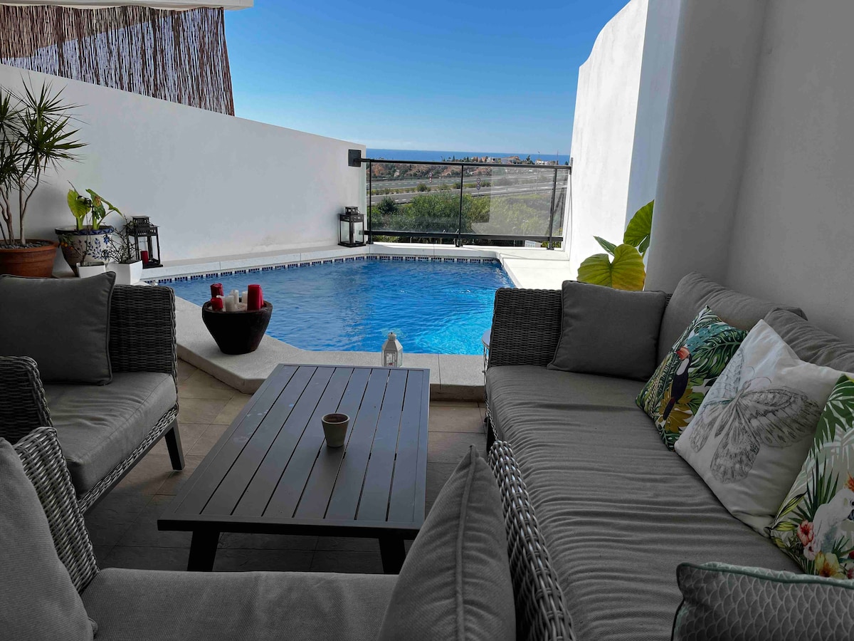 A shaded outdoor seating area is presented, featuring comfortable gray wicker sofas arranged around a dark wooden table. A private pool glimmers in the background, surrounded by a low stone wall. Lush green plants are positioned nearby, adding a touch of nature.