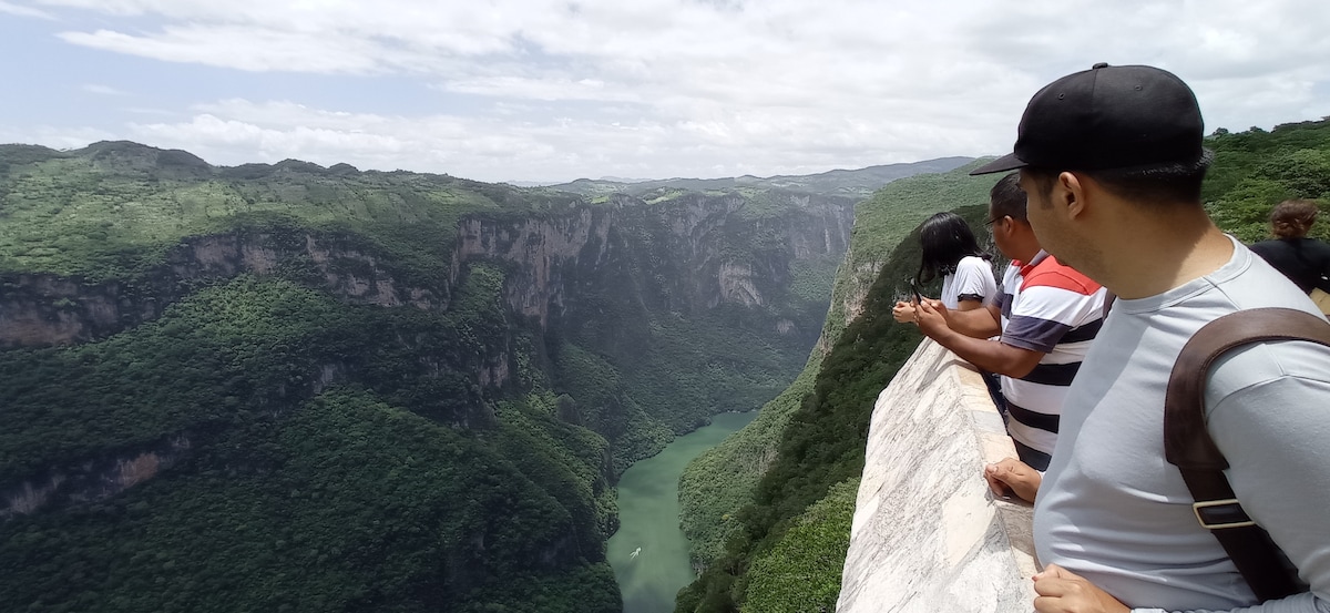 Stunning views of a lush green canyon are captured, with a winding river visible below. Several individuals are positioned along a ledge, admiring the natural beauty of the landscape. The sky is bright with scattered clouds, enhancing the vibrant colors of the surrounding scenery.