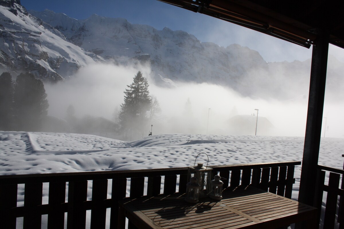 A scenic view is presented from a balcony, featuring snow-covered ground and mist rising across the landscape. Towering mountains serve as a backdrop under a clear blue sky, while a wooden table with lanterns is positioned in the foreground.