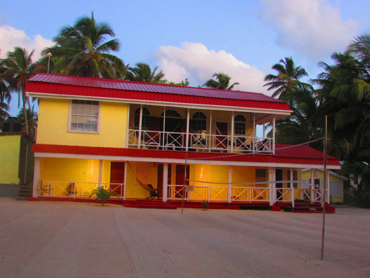 The two-story building features a vibrant yellow exterior with a red roof. A spacious verandah is visible, framed by white railings and adorned with hammocks. Palm trees are scattered around, contributing to the tropical setting. Soft lighting highlights the structure in the evening ambiance.