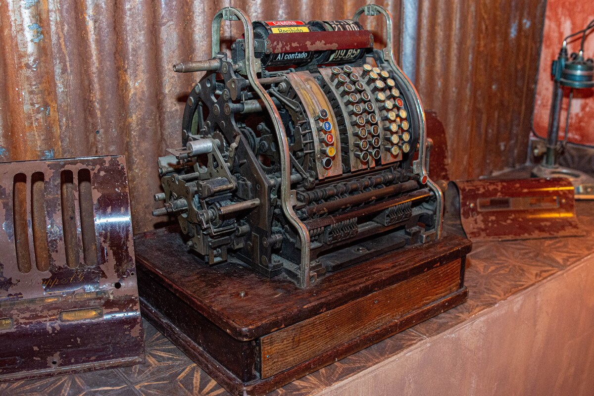 A vintage cash register sits on a wooden base, showcasing intricately designed buttons and levers. The metallic body exhibits signs of age, complementing the textured backdrop of corrugated metal. The scene captures a quaint, nostalgic atmosphere that reflects the historical essence of the space.