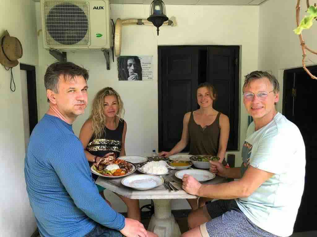 A round marble table is set with several plates of local dishes. Four people are seated around the table in a cozy dining area. An air conditioning unit is mounted on the wall, providing a comfortable atmosphere.