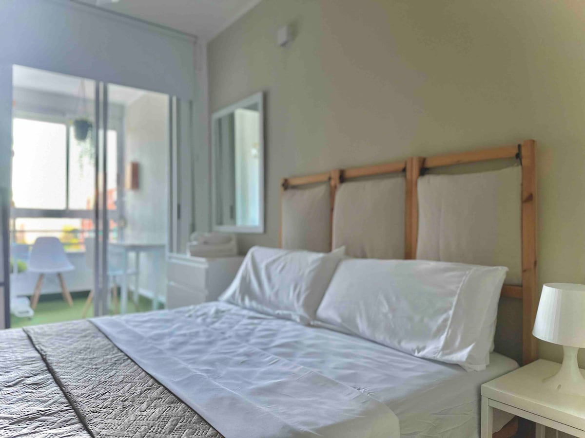 A spacious bed is positioned against a wooden headboard, complemented by crisp white linens. Natural light floods the room through large glass doors, offering views of a private terrace. A mirror and a bedside table are visible, enhancing the room's functional layout.