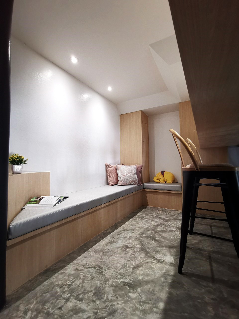 A cozy reading nook is featured, complete with a cushioned bench and decorative pillows. A small plant sits on the side, adding a touch of greenery. Soft lighting illuminates the space, while a modern bar stool is positioned nearby, contributing to the inviting atmosphere.