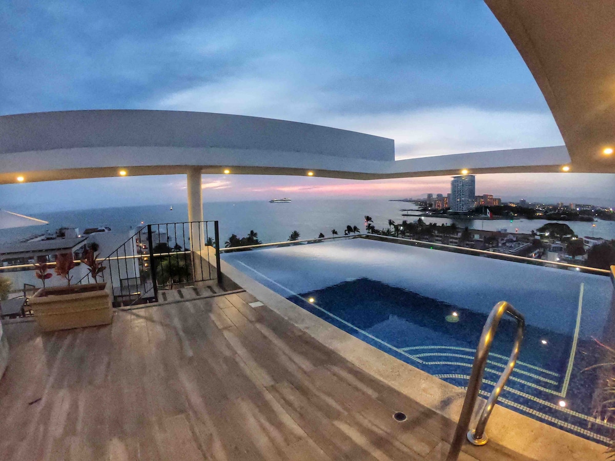 A rooftop area features a swimming pool with a panoramic view of the bay. The horizon is graced with soft evening hues as the sun sets. Lounge chairs are positioned nearby, providing a space to relax while taking in the scenic surroundings.