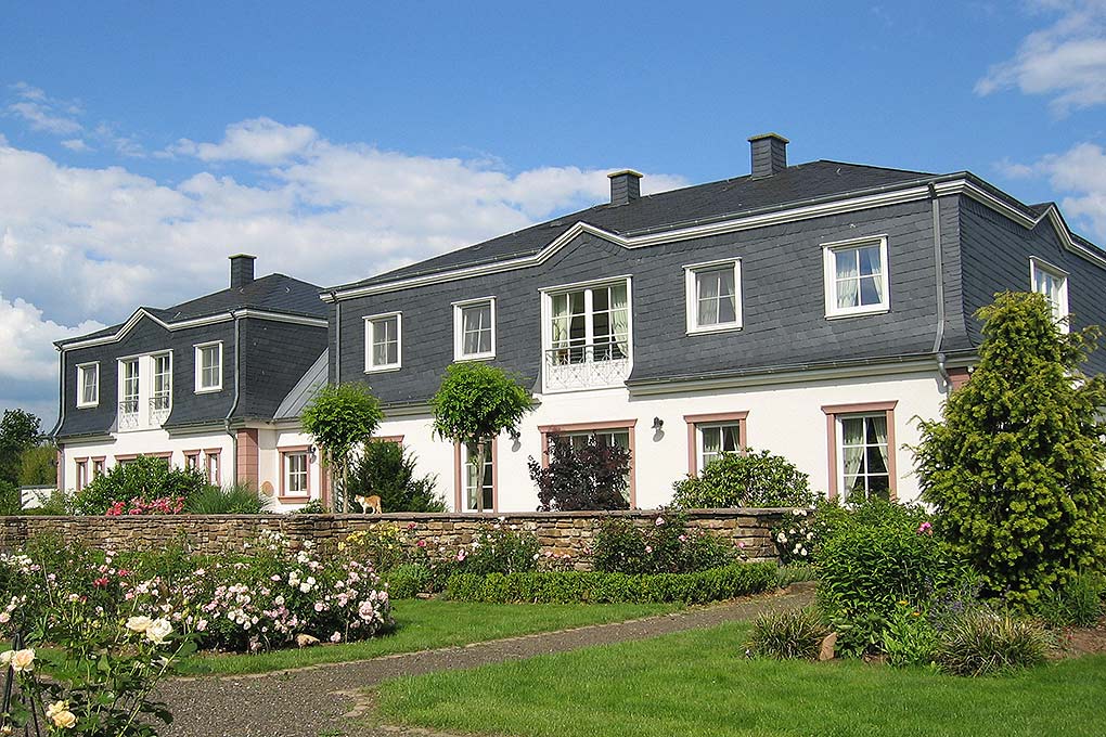 A two-story building features a symmetrical facade with pitched roofs and balconies. Lush gardens with various rose bushes enhance the exterior, while manicured lawns provide open space. The setting presents a covered walkway and vibrant greenery, contributing to a serene atmosphere.