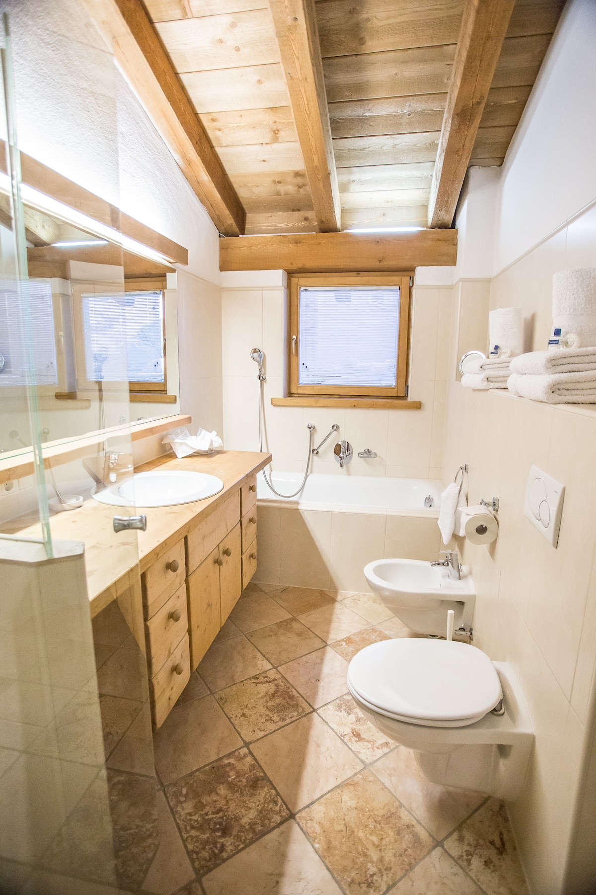 A spacious bathroom features natural wooden beams overhead, enhancing the rustic feel. Two sinks are set within a stone countertop, while a bathtub and shower are situated against the tiled wall. Towels are neatly arranged on the shelving, and a window allows for natural light.