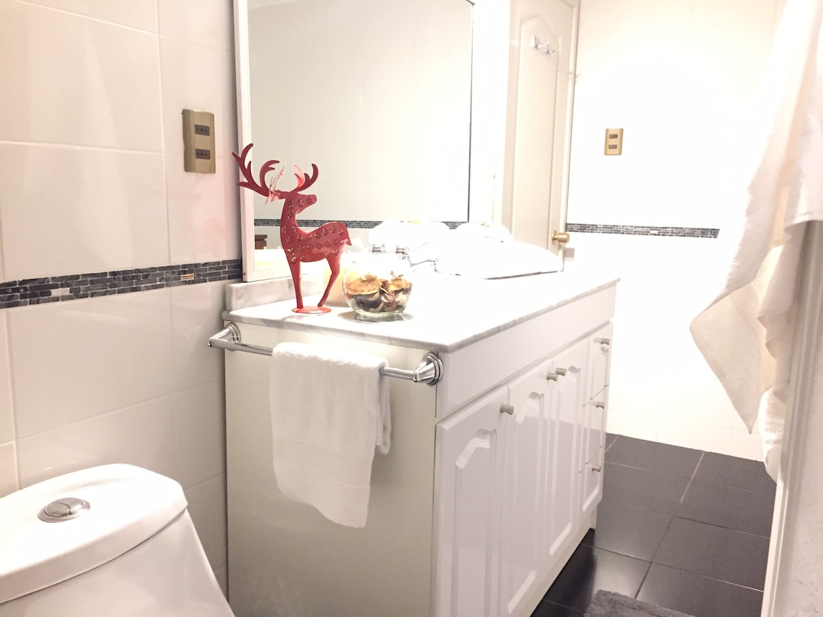 The bathroom features a modern design with white tiles and dark flooring. A double sink vanity is situated against the wall, complemented by a large mirror. Decorative elements include a red reindeer figurine and a glass container with assorted items on the counter. Towels are neatly hung nearby.