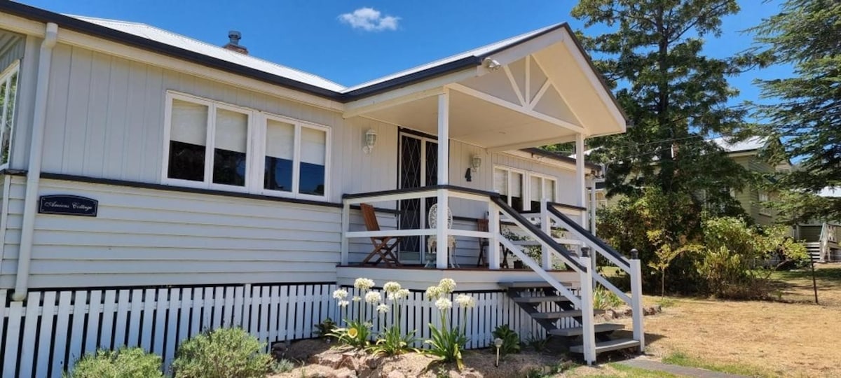 A charming Queenslander cottage features a welcoming front porch with wooden steps leading to the entrance. Two wooden chairs are positioned on the porch, while white flowers and low shrubs enhance the front garden. The exterior showcases classic timber cladding and a gabled roof.