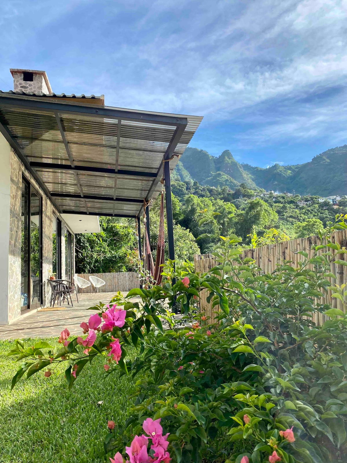 A view of the house from the garden showcases a covered patio with modern architecture. Lush greenery and colorful flowers frame the space, while the backdrop features rolling mountains under a bright sky, creating a tranquil setting for outdoor relaxation.