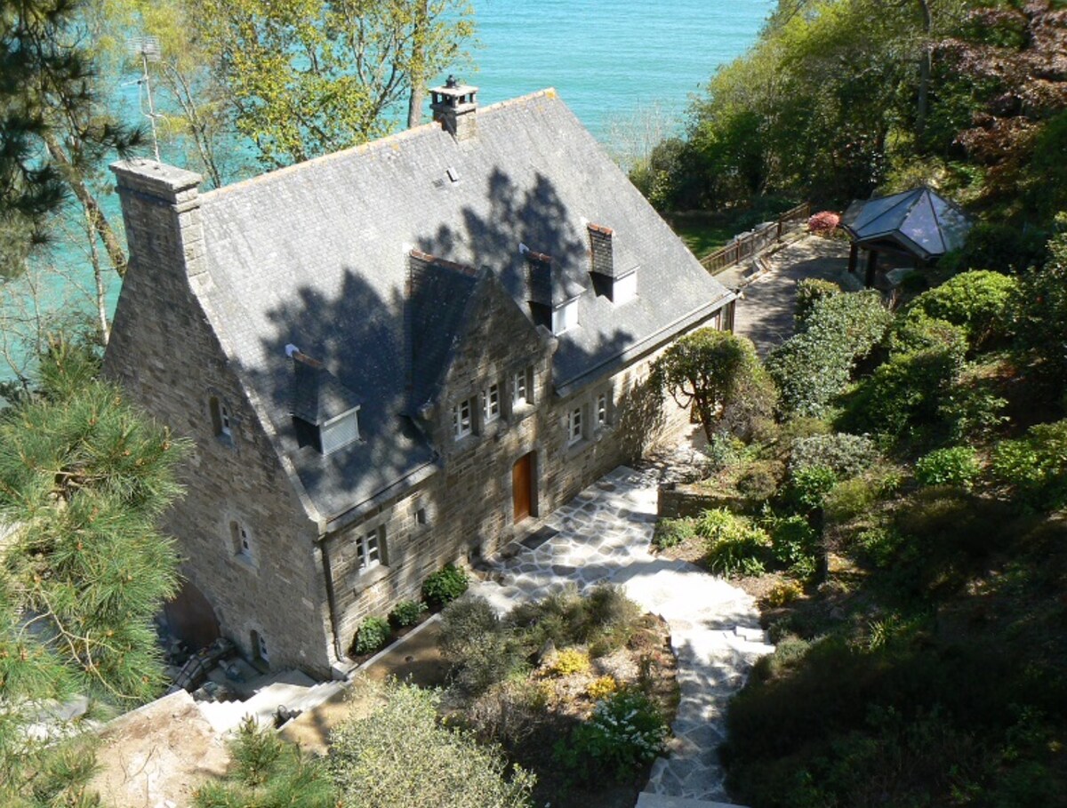 The stone house, surrounded by lush greenery, is situated on a hillside overlooking the sea. A pathway leads down to the tranquil water. The landscape includes various shrubs and trees, enhancing the natural beauty of the coastal environment.
