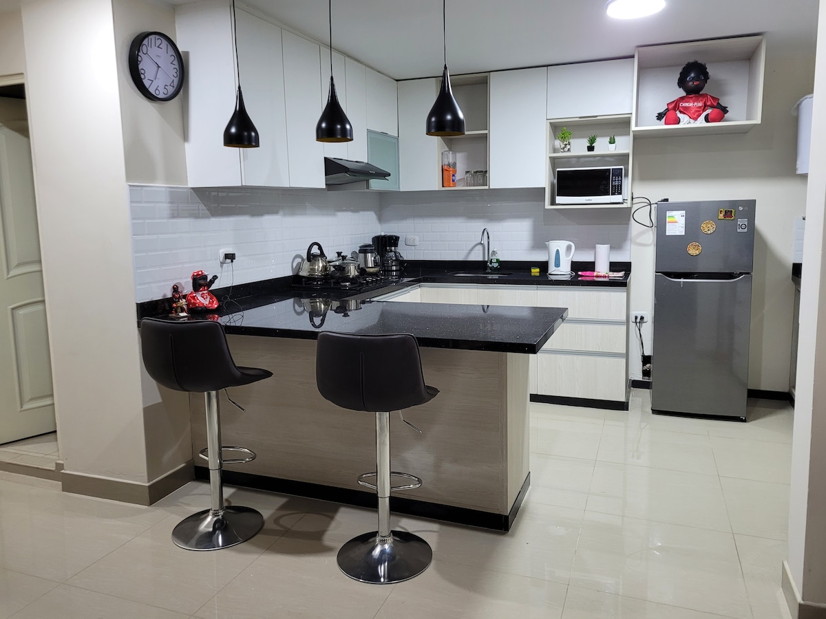 The kitchen features a modern design with a central island and two bar stools. Sleek cabinetry houses essential appliances, including a refrigerator and microwave. Pendant lights hang above the island, illuminating the space, while a clock adorns the wall for easy timekeeping.